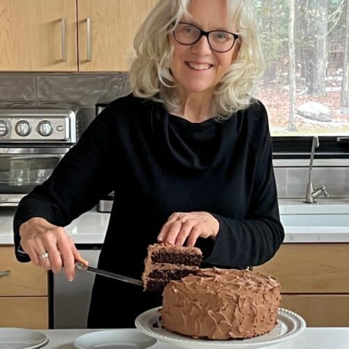 Woman standing behind a kitchen counter, cutting a slice from a chocolate cake.